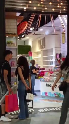 The video shows the entrance to a store called Beauty Plus. Several people are visible, including customers and a cashier with red hair. A young couple is standing near the entrance, embracing and kissing. One of them is holding a pink shopping bag. Other people are walking by or browsing inside the store. The interior of the store is brightly lit with white shelves and product displays.