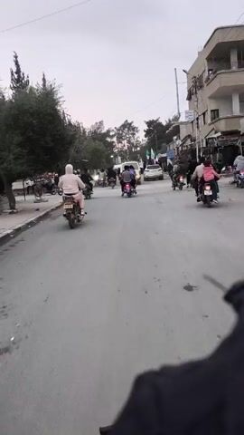 The video shows a group of people riding motorcycles down a street. There are several motorcycles in the foreground and background, and they are all moving in the same direction. A white van is in the middle of the group. A vehicle with a license plate is visible. Some of the riders are wearing helmets. There are buildings on either side of the street. There are flags being carried on some of the motorcycles. The sky is overcast.