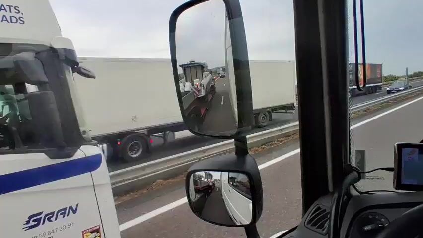 The video shows the interior of a truck cab, looking forward and slightly to the right. The driver is on a highway, with traffic visible in front and in the side mirrors. The truck is carrying a large white boat on a trailer, which is visible in the left side of the frame and in the side mirrors. The boat has "YAMAHA" and "PILOT 8" written on it. The truck is in motion, and other vehicles, including cars and other trucks, are on the highway. The sky is overcast. The side mirrors reflect the highway and surrounding traffic.