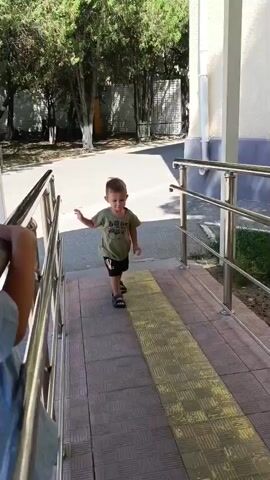 This video shows a young boy walking down a ramp. He is wearing a green t-shirt and black shorts. The ramp has yellow tactile paving and metal handrails. In the background, there are trees, a fence, and a building. The sun is shining, indicating it is daytime. The boy appears to be happy and energetic as he walks down the ramp. The text "baby" is visible on the boy's t-shirt.