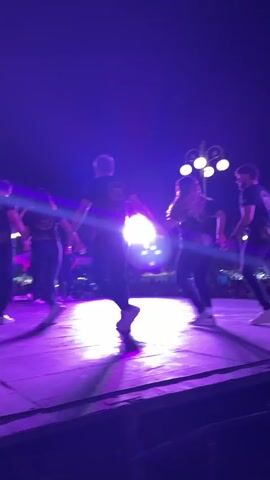A small group of people is on a stage at night, performing some sort of dance in a circle while holding hands. They are under the light of stage lamps that cast a bright purple hue. The dancers are wearing dark pants and T-shirts with logos on them. Outside of the performance space, a crowd of observers are visible. In the background, some decorative lamp posts can be seen.