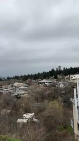 The video shows a landscape view under a cloudy sky. In the mid-ground, there's a cluster of houses nestled on a hillside, interspersed with trees and vegetation. A city skyline can be seen in the distance, partially obscured by trees. The foreground features trees and shrubbery. The video seems to be taken from an elevated position.