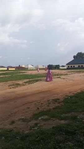 A wide shot captures an outdoor scene with a dirt path in the foreground and a grassy field beyond. Several figures, some wearing pink garments, are scattered across the field. In the background, buildings with white and brown exteriors are visible, along with trees and a cloudy sky. The overall atmosphere suggests a possibly rural or suburban setting.