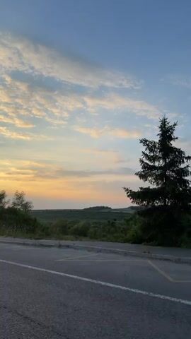 The video shows a moving shot of a paved road with double white lines, taken from the perspective of a vehicle traveling along it. To the left of the road, there is a grassy verge with some trees and bushes. Beyond the verge, a rolling landscape unfolds, characterized by fields and more trees, with a gentle gradient towards the horizon. The sky above is a clear blue with scattered, wispy clouds that are tinged with the warm colors of a sunset, indicating the time of day is late afternoon or early evening. A utility pole with wires is visible on the left side of the frame. The overall scene conveys a sense of peaceful travel through a rural setting at dusk.
