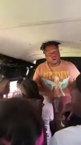 The video shows a man standing in what appears to be the aisle of a bus, singing and dancing. The man has a brown t-shirt and dreadlocks. He raises his hands and dances with enthusiasm. Several other people can be seen sitting in the bus seats. The bus interior is white. It seems like a lively and energetic scene inside the moving bus.