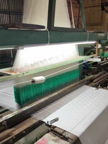 This video shows a close-up view of an industrial loom in operation. The loom is producing white fabric with a subtle patterned texture. Vertical threads of white and green are visible in the upper part of the machine, suggesting the weaving process. A spool of white thread is mounted on the loom. The machinery appears to be old and made of metal, with some wooden components. The lighting is industrial, with fluorescent tubes illuminating the workspace. The overall environment appears to be a factory floor. The video shows no people.