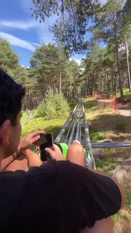 A first-person perspective video shows a person riding a roller coaster through a forest. The person is holding a phone and appears to be recording the ride. The roller coaster track is visible in front of the person, winding through tall pine trees. The sky is blue with some clouds. In a few frames, a second person is seen with their arm around the rider, and they briefly lean in towards the rider's face.