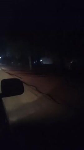The video shows a view from inside a car, likely from the passenger seat, driving down a dark road at night.  Streetlights illuminate the road ahead. A car is visible in the distance, moving in the same direction. The car's side mirror is visible in the lower left of the frame. The road is mostly clear and has a reddish hue under the streetlights. The surrounding area appears dark and indistinct, possibly with trees or bushes lining the roadside.
