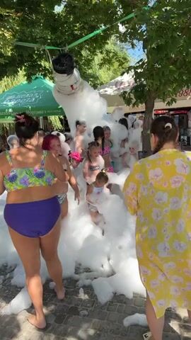 A foam party is taking place outdoors with a foam machine in the background. Children and adults are immersed in the foam, some playing, others watching. The event appears to be at a resort or a public area with trees and tents providing shade.