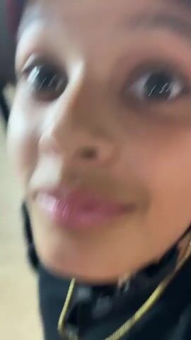 A close-up video shows a young person wearing a red, black, and white helmet with graphics. The person is facing the camera and then turns to the side, sticking out their tongue. The video is shaky, suggesting it was filmed while in motion. The person is wearing a black t-shirt and a gold chain. The background appears to be an indoor setting, possibly a skate park or similar recreational area, with neutral-colored surfaces. The person's expression changes from neutral to playful.