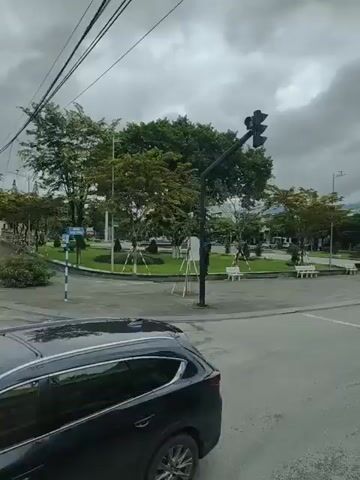 The video shows a street scene under a cloudy sky. A traffic light is visible in the foreground. A green car drives by, followed by motorcycles. A white SUV with red rims is parked on the side of the road. The scene appears to be a public space with trees, benches, and a grassy area.