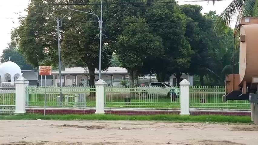 The video shows a scene with a white truck parked behind a green fence with white posts. In the background, there is a building with a white dome and a sign that says "CALLING". There are also several trees and a brown tank on the right side of the frame. The ground in the foreground is dirt. There are several animals, possibly goats, walking behind the fence in the middle ground. The overall impression is that of a rural or semi-rural environment, possibly a train station or a religious site.