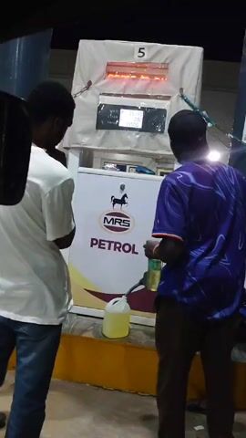 Three people are standing at a gas pump at an MRS Petrol station. One person is wearing a white t-shirt and jeans and faces the pump. A second person is wearing a blue and purple t-shirt, brown pants, and a black baseball cap. He holds a small bottle in his right hand and is facing the pump. The third person is barely visible, standing between the other two. The gas pump has a digital display showing the amount of petrol dispensed and the price. A yellow container sits on the ground next to the pump. A blue basket and a black bucket are visible to the right of the gas pump, next to a large metal cylinder. The scene appears to be taking place at night.