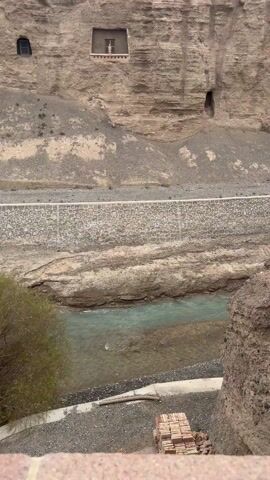 The video shows a small, light blue river running through a rocky terrain. The river is bordered by a retaining wall made of irregular stones. Above the retaining wall is a steep, rocky hillside with cave-like openings cut into it. The hillside is mostly barren with sparse vegetation. The camera pans upwards and then back down, focusing on the river and the hillside. There are also stacks of bricks visible on the lower right side of the frame. The sky is overcast.