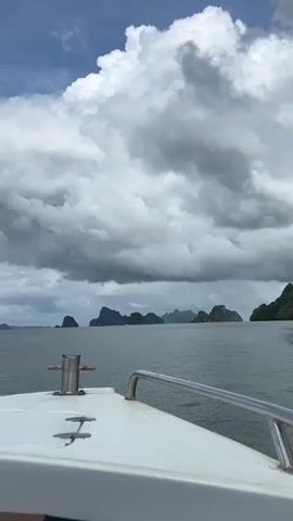 The video shows the bow of a white boat moving through choppy gray water. In the distance, several large, green islands with steep, rocky cliffs are visible under a sky filled with dramatic, dark gray clouds. The sun is partially visible through breaks in the clouds, casting light on the water and highlighting the boat's chrome railing and a silver mooring cleat. The overall impression is of a journey through a rugged, tropical seascape with a stormy sky.