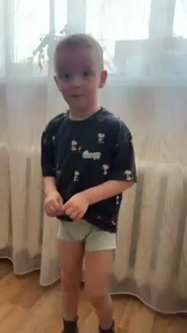 A small child stands indoors near a window. The child has short blonde hair and is wearing a black t-shirt with an animated character print and light green shorts. The child is smiling and appears to be speaking. Behind the child is a white curtain, a window with plants, and a radiator. The floor appears to be made of wood.