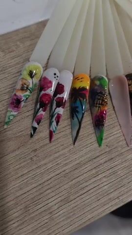A shallow depth of field shot captures a fan of ten artificial nails fanned out on a wooden surface. Each nail is displayed on a white plastic holder and features a unique design. The nails are arranged in a curve, with the tips pointing towards the right. The designs include floral patterns, a tropical sunset scene with palm trees, a leopard print with gold accents, and a butterfly graphic. The lighting is soft, illuminating the intricate details of the nail art. The wooden surface exhibits a natural grain pattern. The background is blurred, focusing attention on the main subject.
