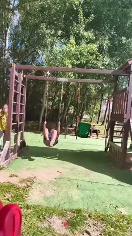 A young boy on a swing set is swinging back and forth. He is wearing a blue shirt and shorts. To the left of the swing set, another child is standing and watching. In the background, there is a wooden play structure with a slide and a green tire swing. The area is covered in artificial turf, and there are trees and foliage in the background. The sun is shining.