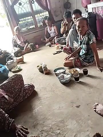 A group of people are sitting on the floor of what appears to be a home. There are several bowls of food and drinks on the floor in front of them. The room has a window with curtains and a wooden door. Some of the people are looking at the camera, while others are looking at each other or at the food. One person is holding a phone. The floor is made of concrete.
