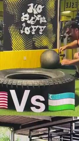 The video depicts a man flipping a large tire. He is wearing a tank top and black shorts. The man begins by placing his hands on the tire, then he lifts it to a standing position. He then pushes the tire over, causing it to fall to the ground. He then repeats the process. The background includes a green artificial turf floor, a wall with a yellow and black design, and exercise equipment. The video contains graphics showing the United States and Uzbekistan flags with the word "vs" between them.