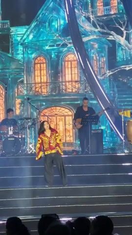 A female singer, dressed in a red and yellow patterned top and dark trousers, performs on stage with a live band. The band consists of a drummer on the left, a bassist to the right of the singer, a guitarist further right, and another musician playing percussion instruments like congas on the far right. They are positioned on a multi-tiered stage.

Behind the performers, a large LED screen displays a striking backdrop. Initially, it shows a stylized, ornate "ice house" or crystalline building structure in shades of blue with warm orange glows emanating from its windows. As the camera zooms out, the screen above the building illuminates with large yellow text. The text reads "MANG ĐEN" and "KHÁT VỌNG VƯƠN XA," indicating the event is taking place in Măng Đen, Vietnam, with "Khát Vọng Vươn Xa" translating to "Aspiration to Reach Far."

Bright spotlights and blue stage lights illuminate the scene, creating a dynamic concert atmosphere at night. The heads of audience members are visible in the foreground, watching the performance.