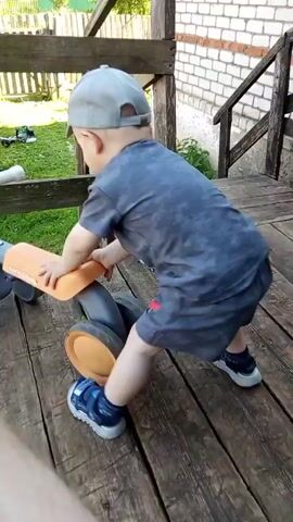 A child wearing a grey cap, a blue tie-dye t-shirt, grey shorts, and blue sneakers is playing with a grey and orange balance bike. The child is facing away from the camera, with their hands on the handlebars and their legs on the seat, as if trying to ride the bike. The child is on a wooden deck outdoors, with a wooden railing in the background. The deck appears to be part of a porch or playground. Green grass and foliage are visible beyond the railing. The camera is focused on the child and the balance bike, with a blurred foreground element that appears to be a person's knee. The lighting suggests it is daytime.