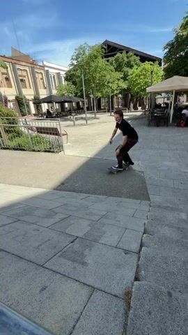 A young man with blonde hair and tattoos on his arms is skateboarding down a sloped concrete path on a sunny day. He is wearing a black t-shirt and brown pants. He is in the process of performing a trick as he descends. The surrounding environment includes buildings with windows and balconies, trees with green leaves, and a metal railing in the foreground. There are also tables and umbrellas visible in the background, suggesting an outdoor seating area. The sky is clear and blue.