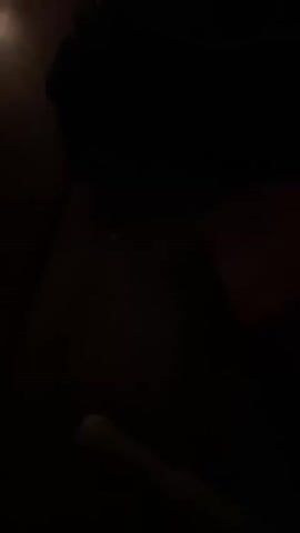 The video shows a close-up of two dark metal bowls on a wooden surface. A hand with red nail polish is seen striking one of the bowls with a wooden mallet, producing a sound. The scene is dimly lit, with a single lit candle visible in the background, casting a warm glow on the wooden surface. The remainder of the environment is obscured by darkness.