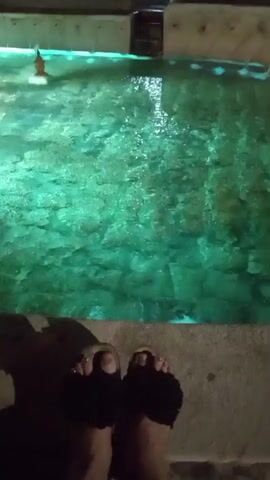The video shows a person's feet wearing flip-flops standing at the edge of a swimming pool. The pool is lit with green underwater lights, and the water appears clear, with visible stones at the bottom. There is a small, floating object in the pool, possibly a toy or a light. The environment appears to be outdoors, possibly at night, given the lighting. The person is not in the water, but rather standing on the pool's edge.