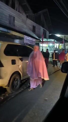At night, a woman in a purple hijab walks past a white car parked on the side of a street. Behind the car, several men are walking down the street. The buildings on the left are made of wood and have a rustic appearance. There are signs and flags visible on the buildings. The street is dark, with some artificial lighting from the buildings and possibly from the camera's flash. The scene appears to be in a residential or commercial area.