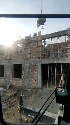 The video shows a construction site with a partially built brick building. A crane is visible in the frame, carrying a container or load towards the building. The building appears to be multiple stories high, and scaffolding is set up around it. There is a person visible on one of the upper levels of the building. The ground around the building is muddy and cluttered with construction debris. The sky is partly cloudy with the sun shining brightly.