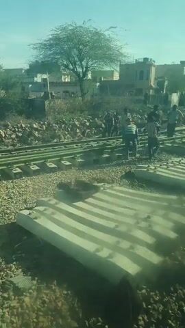 The camera is on board a moving train, filming the surroundings as it passes by.  A small group of people are visible near the train tracks, seemingly involved in railroad construction or maintenance. Several concrete railway ties are positioned on the gravel bed adjacent to the tracks, ready to be installed. The setting appears to be a rural or suburban area, with scattered buildings and sparse vegetation dotting the landscape. The weather seems clear, with a blue sky visible above.