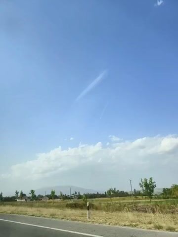 The video captures a clear blue sky with scattered clouds and two contrails. Below the sky, a flat landscape is visible, featuring dry grass, a road with white markings, a few trees, and some houses. In the distance, mountains can be seen. The sun appears to be high in the sky, suggesting daytime. The camera is in motion, likely from a vehicle, as indicated by the movement of the road and landscape. There are no people or animals visible in the frame.