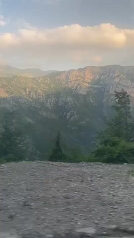 The video shows a rocky, elevated landscape with mountains in the background. The sky is blue with some white and pink clouds. The mountains are steep and covered with green vegetation, with some bare rock faces visible. The foreground is a gravelly surface with some sparse greenery. There are no people or animals visible. The video appears to be taken from a moving vehicle due to slight blurriness and motion. The lighting suggests it is either early morning or late afternoon.