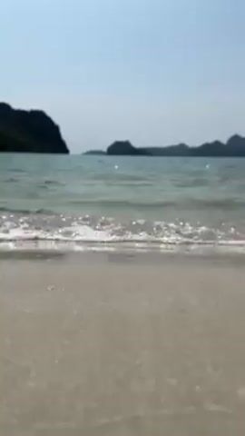 The video depicts a tranquil beach scene with gentle waves lapping onto the shore. The sand is light in color. In the background, there are islands with dark green vegetation visible against a clear, light blue sky. The focus is on the water meeting the sand, with small bubbles visible in the wave crests.