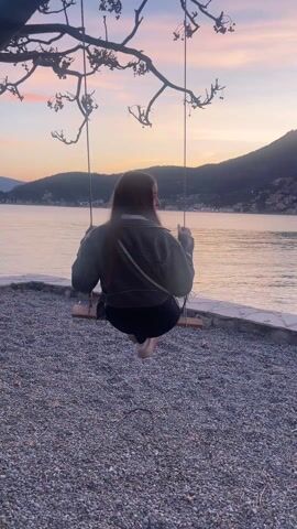 A young woman sits on a wooden swing suspended from a tree branch, overlooking a body of water. She is facing away from the camera, then turns to look over her shoulder with a smile before returning to facing forward. She is wearing a green jacket, black leggings, and light-colored shoes. She also has a small white bag slung across her body. The ground is covered in small pebbles. In the background, there is a body of water, with land and a mountain in the distance, and a pink and orange sky indicating it is sunset or sunrise.