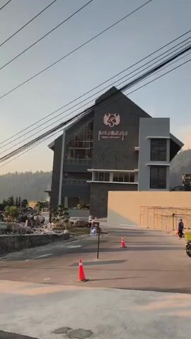 A video captures a street scene at what appears to be either dawn or dusk. The camera pans from a building with scaffolding and construction materials in the foreground, past a stone wall, to a multi-story house with a circular balcony and a tall evergreen tree. A purple truck is seen driving down the road towards the camera. The sky is clear with a gradient of colors suggesting sunrise or sunset. Power lines are visible throughout the shot.
