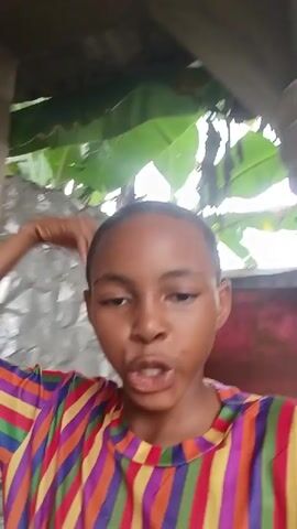 The video shows a young person wearing a brightly striped shirt, looking directly at the camera. Their mouth is open as if speaking, and their right hand is gesturing downwards. The background appears to be outdoors, with large green leaves visible behind them, possibly banana leaves. There is a textured wall or structure to the right and a corrugated surface on the left. The lighting suggests it is daytime.