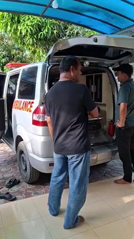 The video shows an ambulance parked under a blue awning, with its rear doors open. Three men are standing near the ambulance, one in a black shirt and jeans, another in a green shirt and black pants, and a third in a white shirt and black pants. A woman in a gray hijab walks by in the foreground. The ambulance has the word "AMBULANCE" printed on its side, with a red cross. Inside the ambulance, there is a stretcher and a fire extinguisher. There are sandals on the tiled ground near the ambulance. The environment appears to be a residential area with trees visible in the background.