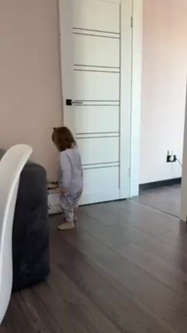 A small child in light-colored pajamas walks on a hardwood floor towards a white door. The child turns towards the camera briefly and then reaches for a bag on the floor near the door. The room has light-colored walls, a grey sofa, and electrical outlets visible near the doorway.