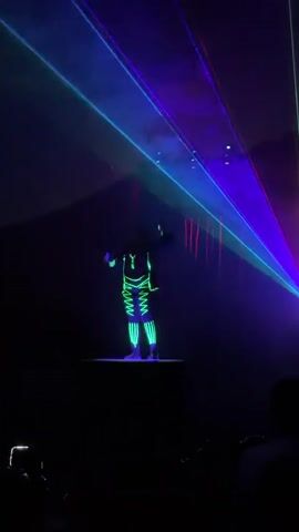 A person dressed in a neon green outfit is standing on a platform in a dark room. They are illuminated by colorful laser lights that are projected from the upper right corner of the frame, creating a dramatic effect. The laser lights form beams of purple, blue, and green, which are reflected on a dark surface above the person. In the lower portion of the frame, silhouettes of people are visible, suggesting an audience. One person in the audience is holding up a phone, likely to record the performance. The overall atmosphere is one of a live performance or show, with a focus on visual spectacle.