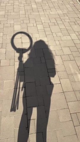 A person's shadow is cast on a sidewalk. The shadow includes the person holding a ring light and stand. The sidewalk is made of light-colored rectangular paving stones. The person is wearing sneakers.