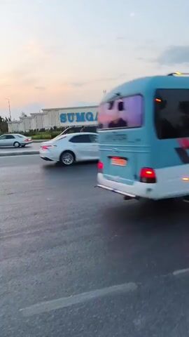 A street scene during twilight with a building displaying the illuminated sign "SUMQAYIT". Several cars are on the road, including a white SUV, a black sedan, and a black minivan. A pedestrian is crossing the street. The sky is a blend of blue and pink hues, indicative of sunset.