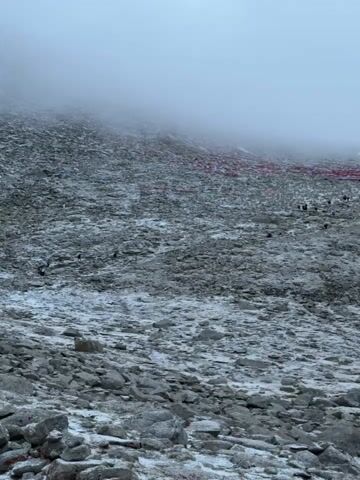 The video shows a wide shot of a rugged, rocky mountain slope, partially covered in patches of snow. The terrain is dominated by various shades of grey stones and boulders. A thick mist or low-hanging cloud obscures the upper portion of the mountain, creating a diffused, pale sky.

A line of people, appearing small against the vast landscape, can be seen walking slowly up the slope from the lower right towards the upper left. They are scattered across the rocky path, some closer and clearer, others more distant and less defined.

Stretching across the middle distance of the slope, a dense array of colorful prayer flags is visible. These rectangular cloths, in hues of red, blue, yellow, green, and white, are strung horizontally, creating a vibrant band against the muted rock and mist.

The overall atmosphere is one of a challenging, possibly spiritual, journey in a cold, high-altitude environment, indicative of a pilgrimage or a strenuous hike. No specific text is visible in the video.