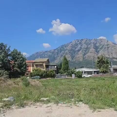 The video pans across a rural landscape under a bright blue sky with scattered white clouds. In the foreground, there is overgrown grass and some debris. In the midground, there are several buildings, including what appears to be a small market or restaurant with white awnings and vehicles parked nearby. Further back, a large, rocky mountain dominates the horizon. The video moves from left to right, revealing more of the buildings and the expanse of sky and mountains.