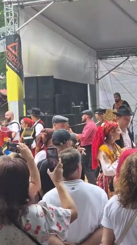 A busy outdoor festival is underway with a band performing on a raised stage. The band consists of several musicians dressed in traditional folk costumes, including vests, hats, and some with red accents. One musician plays an accordion, another a guitar. Several people in the audience are holding up their phones to record the performance. The audience is a mix of ages and appears to be enjoying the event. In the background, there are tents and banners, indicating a marketplace or fair. The overall atmosphere is lively and festive.