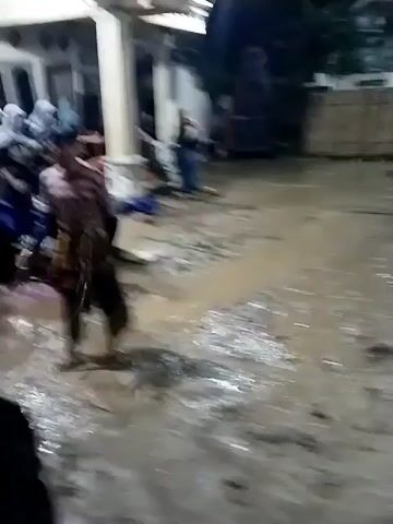 The video captures a nighttime outdoor scene in Jenggrik, Indonesia, where a gathering is taking place on a muddy ground area in front of buildings. The ground is wet and covered in puddles, suggesting recent rain or standing water.

The central activity involves two young men performing. One man, wearing a black long-sleeved shirt and black head covering, holds what appears to be a black rope or whip. He moves around, seemingly interacting with another man wearing a light-colored striped shirt and dark pants, who is dancing or performing dynamic movements. Other individuals, both men and women, are visible in the background, observing the event. Some are standing near the buildings, which are illuminated by artificial lights. One person in the foreground, wearing a black hoodie and a black face mask, briefly looks towards the camera before turning away to watch the performance. The scene conveys a cultural or community event taking place under challenging ground conditions.