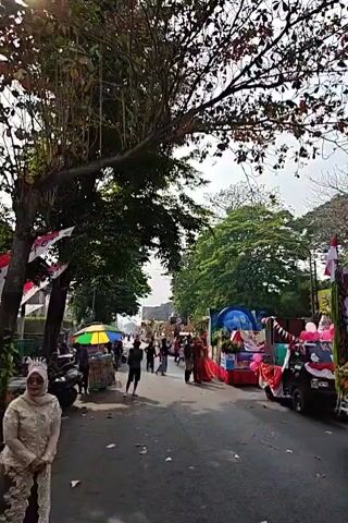 The video captures a street scene, likely during a parade or festival. In the foreground to the left, a woman in a white traditional outfit stands facing the camera, with a tree and foliage above her. The street itself is visible, with various decorated floats or vehicles moving along it. One float is prominent on the right, adorned with fruits, vegetables, and fabric. People are visible walking and standing around the floats, suggesting a public gathering. The environment appears to be outdoors, with trees and buildings lining the street. Several Indonesian flags are also visible, indicating a possible national celebration. The overall atmosphere seems festive and crowded.