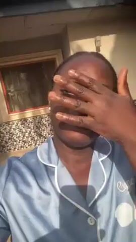 A medium shot shows a dark-skinned woman with hair in small braids, wearing a light blue pajama top, standing outside in front of a wall. She touches her face and gestures with her hands, then makes a kissing face and smiles at the camera. A window with a red frame is visible in the background. The lighting suggests it is daytime.