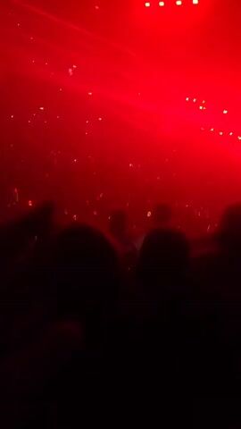 The video shows a concert viewed from the audience. The stage has a large screen with changing graphics, and bright red lasers are sweeping across the crowd. The venue is dark, with the only light coming from the stage and scattered cell phone screens held by members of the audience. People are mostly visible as silhouettes, facing the stage, some with their hands raised. The atmosphere is filled with fog or haze, enhancing the visual effects.